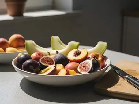 Taça com fruta de verão — pêssegos, ameixas, figos e melão — sobre uma bancada, ao lado de uma tábua.